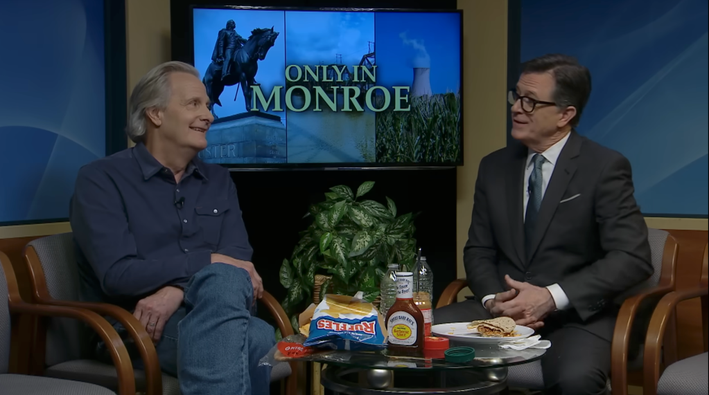 Stephen Colbert and a guest sitting on a talk show set with the words "Only in Monroe" on the screen behind them.