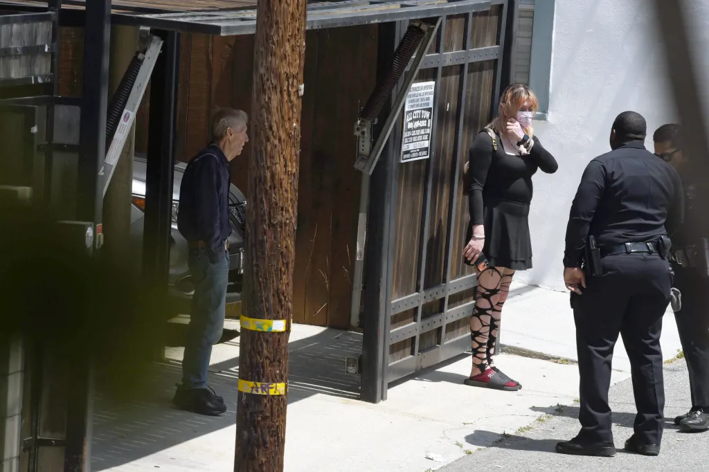 A man, a woman in a mask, and two police officers stand outside a building next to a wooden pole and a sign for "All City Tow".