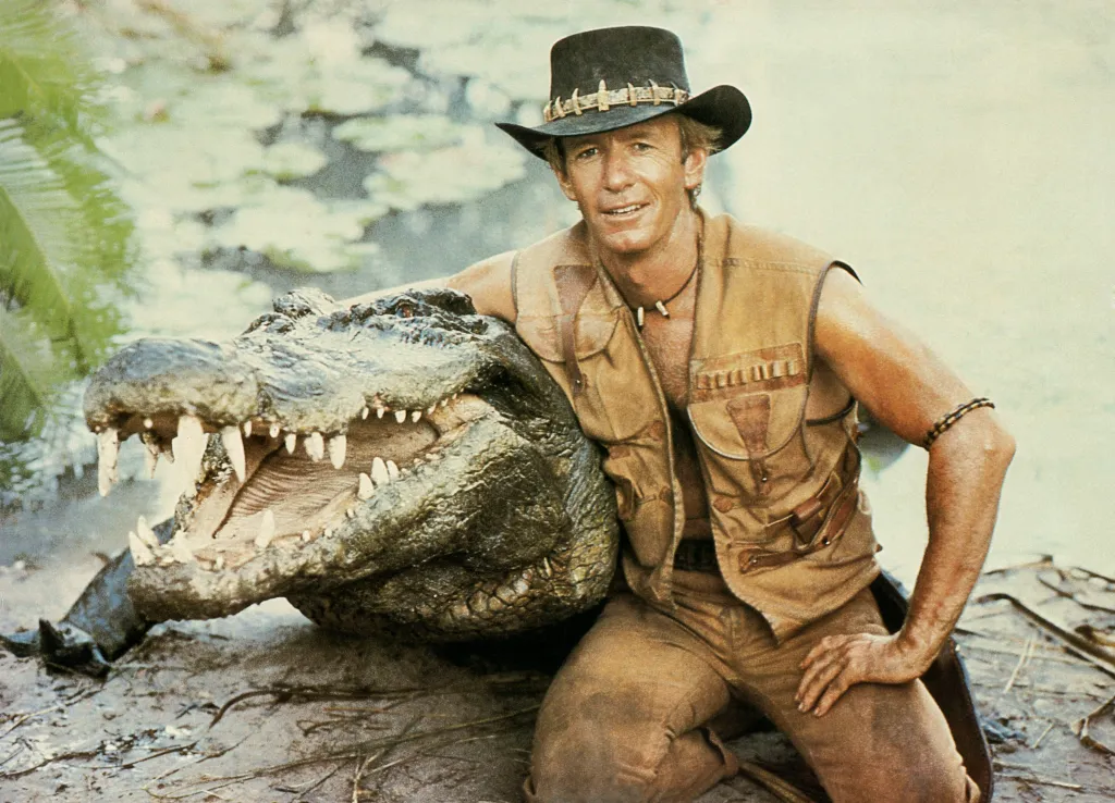 Paul Hogan in "Crocodile Dundee" sitting next to a crocodile with its mouth open.