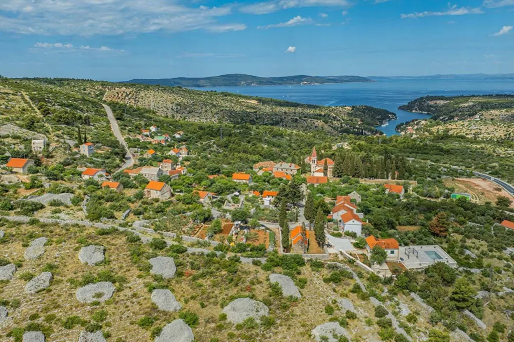 Aerial view of a Croatian village in Dalmatia, with red-roofed buildings nestled among green trees, leading down to the blue Adriatic Sea.