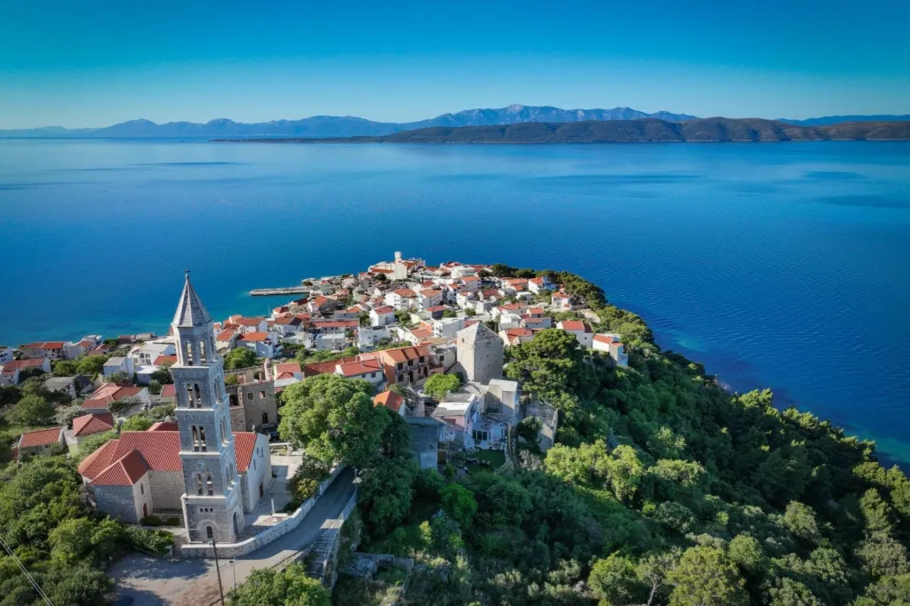 Aerial view of the village of Veli Lošinj on a peninsula, with a large bell tower, red-roofed buildings, and the Adriatic Sea.