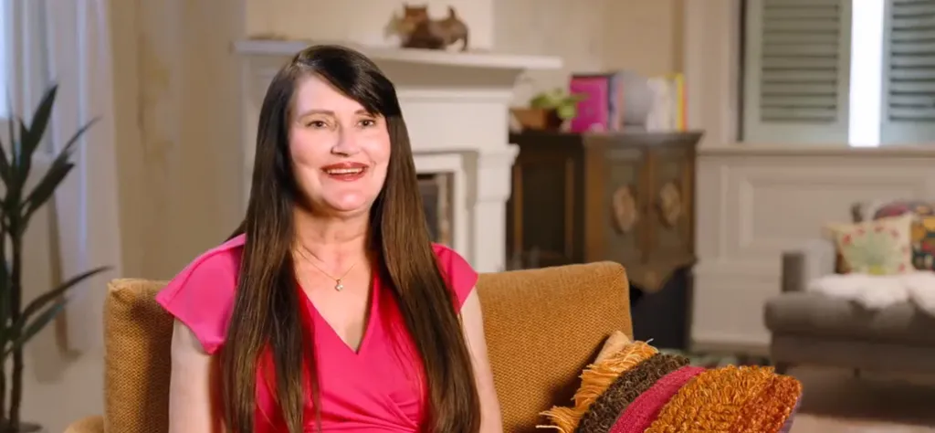 A woman with long dark hair and a pink top smiles while sitting on a brown chair.