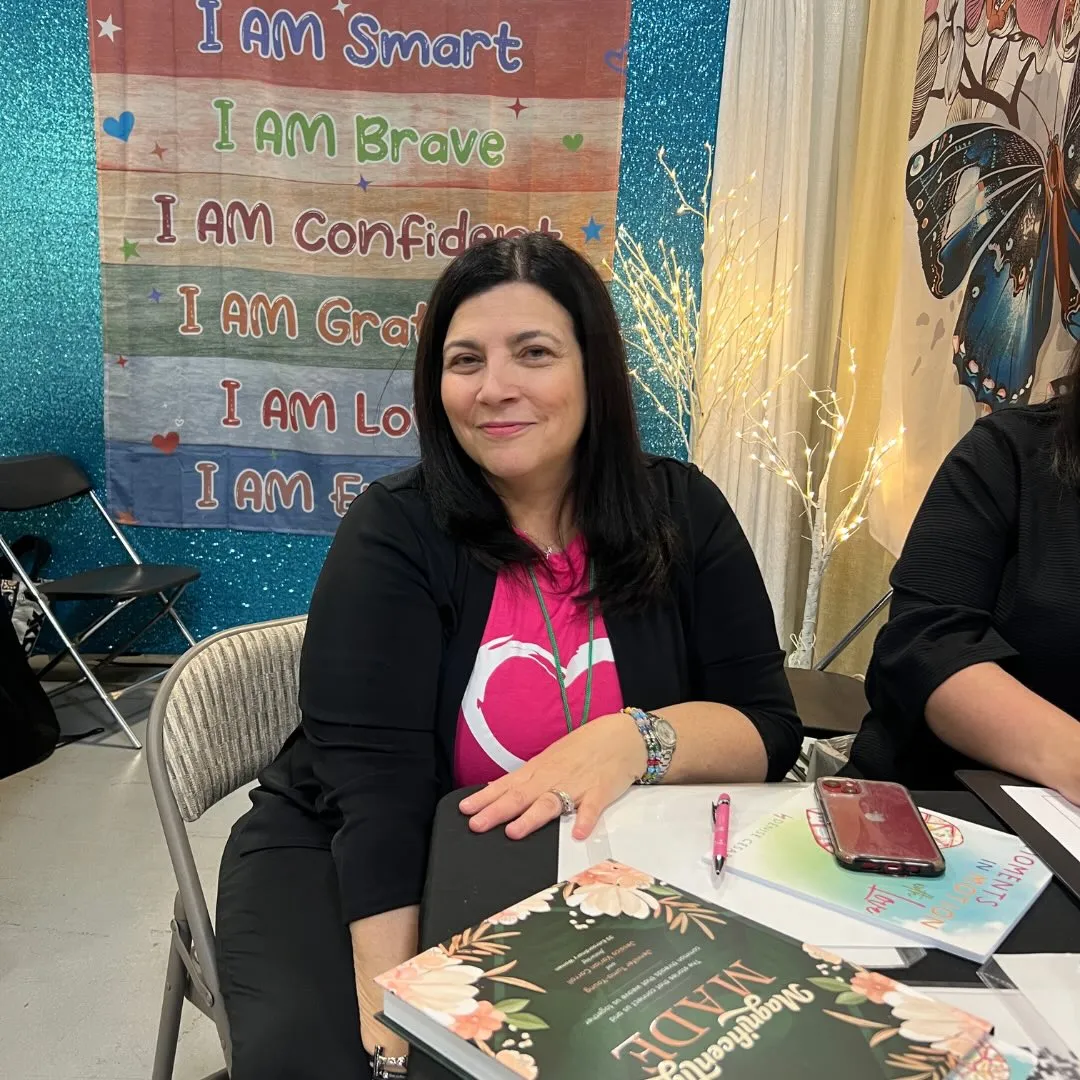 Denise Cesare smiling while sitting at a table with books, with a banner behind her that reads "I am Smart, I am Brave, I am Confident, I am Grateful, I am Loved, I am Enough."