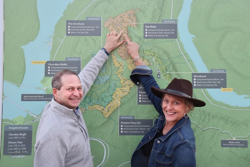 Two people smile at the camera while pointing at a development map with areas labeled "The Overlook," "Big Ridge," "Cherokee Bluffs," and "Pioneer Pass."