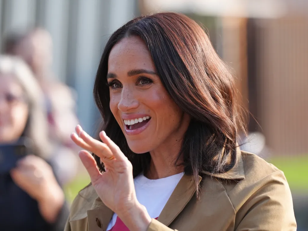 Meghan Markle smiles, holding up her hand as if waving.