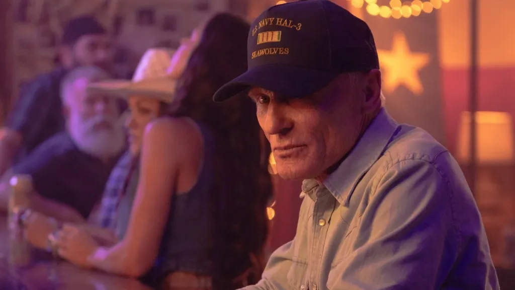 An older man sits at a bar or table in a warmly lit honky-tonk, wearing a navy blue cap reading 