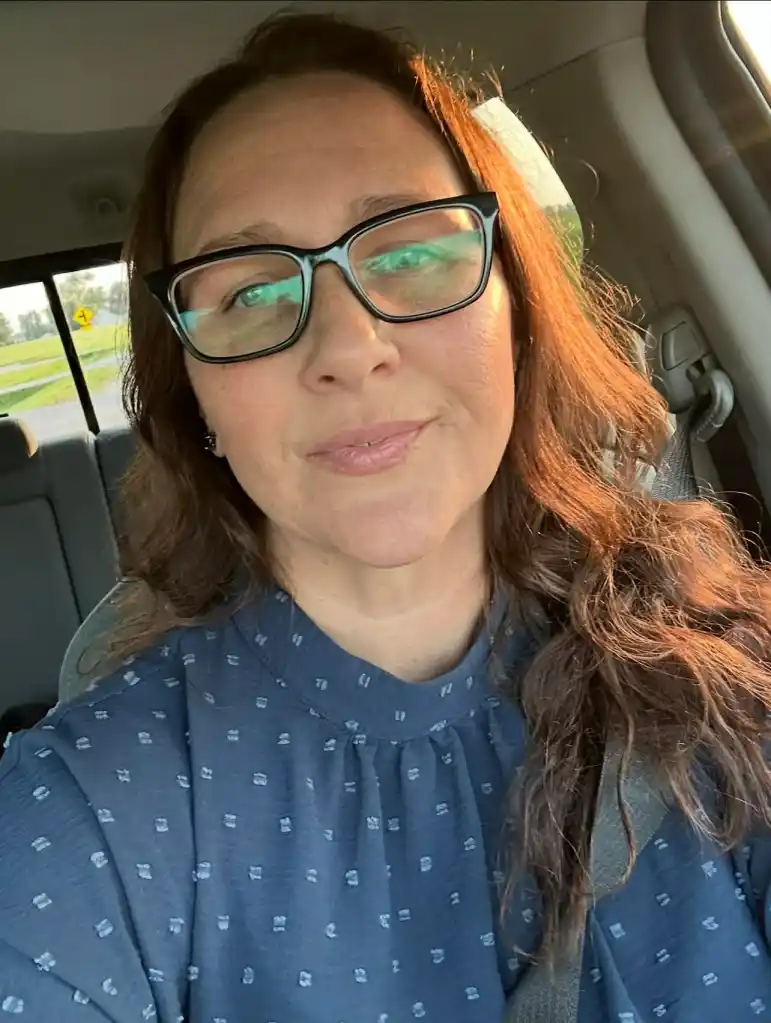 A woman with long brown hair and glasses looks forward in a car