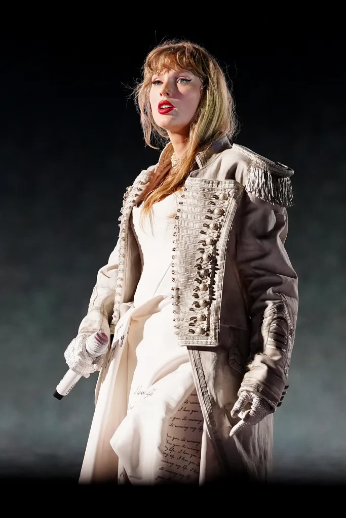 Taylor Swift performing during "The Eras Tour" in a white dress and embellished jacket, holding a microphone.
