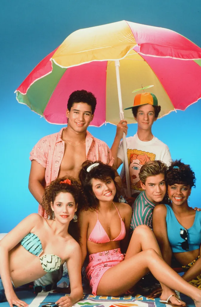 The cast of Saved by the Bell, including Elizabeth Berkley, Mario Lopez, Tiffani Thiessen, Dustin Diamond, Mark-Paul Gosselaar, and Lark Voorhies, posing for a beach-themed photo.