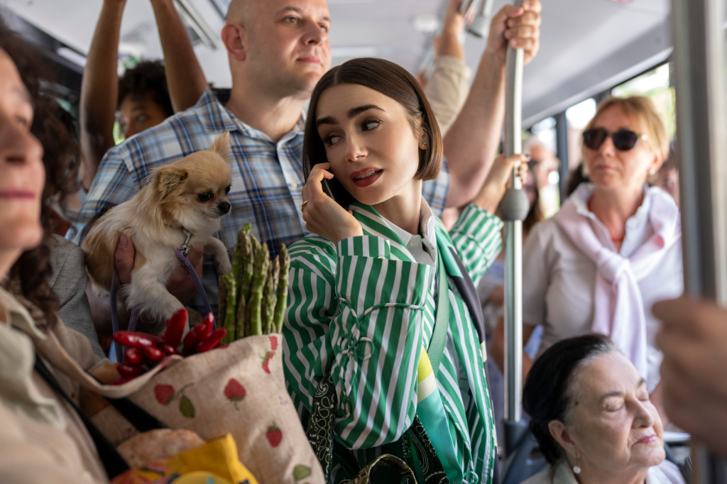 Lily Collins as Emily on a crowded bus, talking on her phone, with a small dog and groceries beside her.