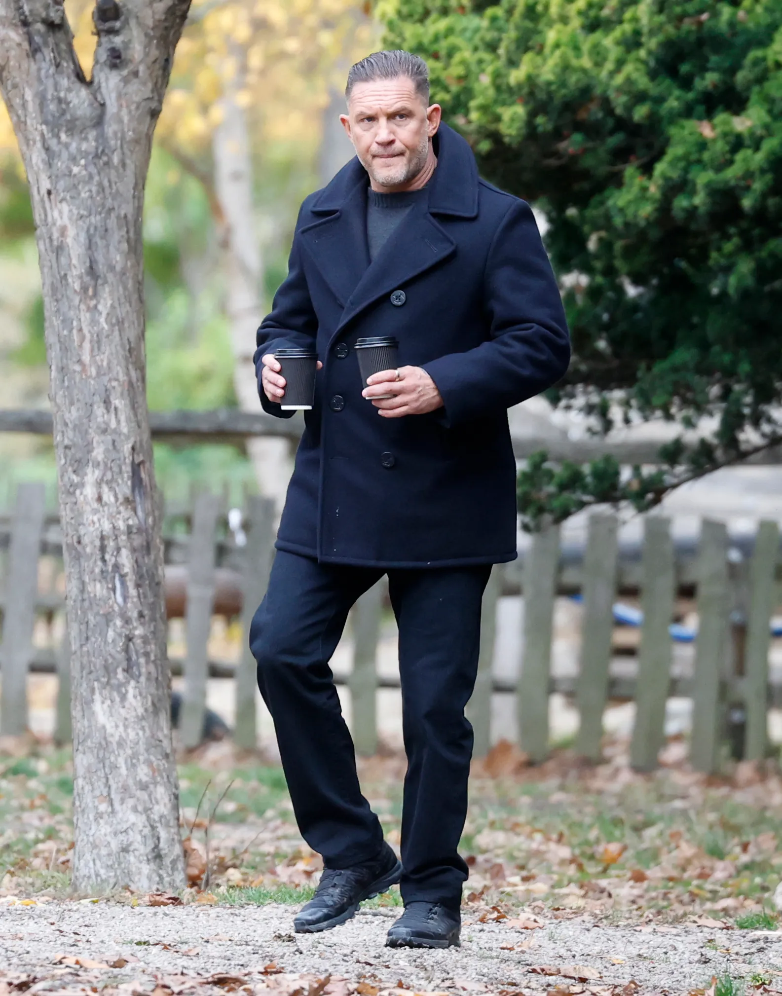 Tom Hardy, dressed in a dark coat and trousers, walks with two cups of coffee.