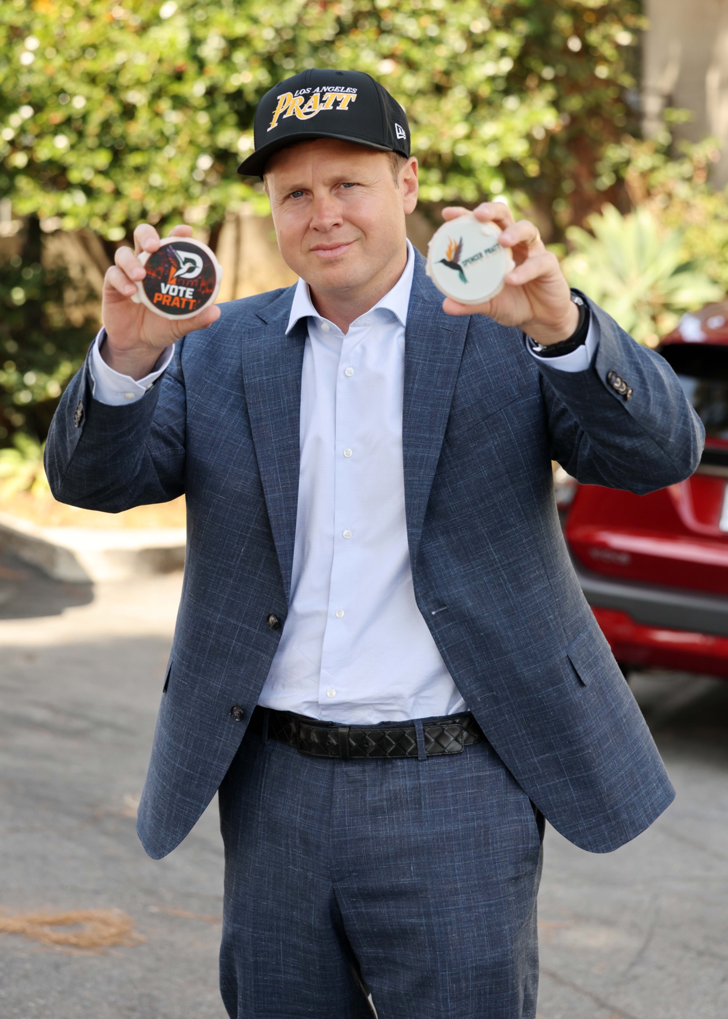Spencer Pratt in a blue suit and black baseball cap, holding two "VOTE PRATT" cookies.