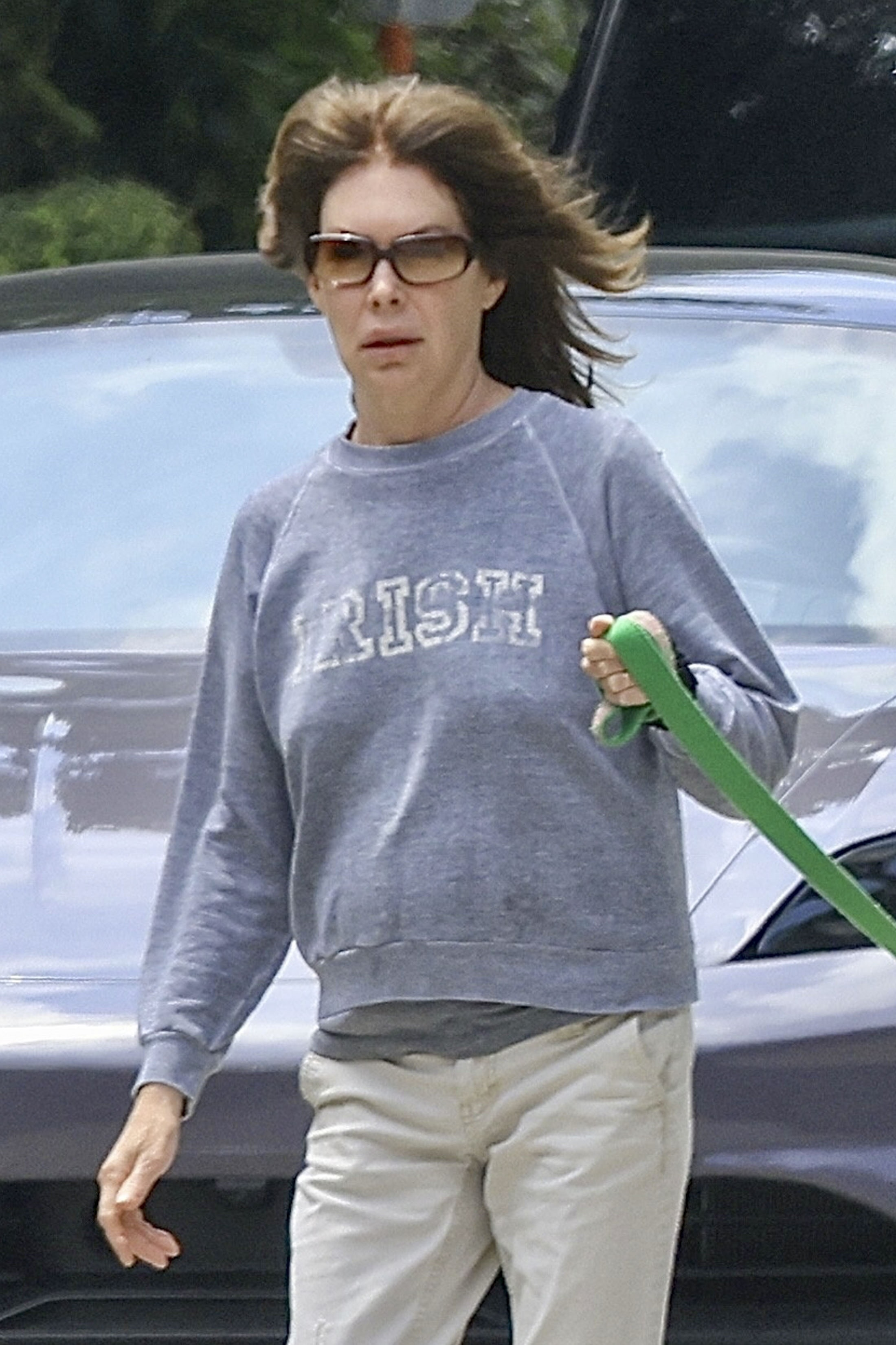 Lara Flynn Boyle in sunglasses, a gray "IRISH" sweatshirt, and khaki pants, holding a green dog leash.