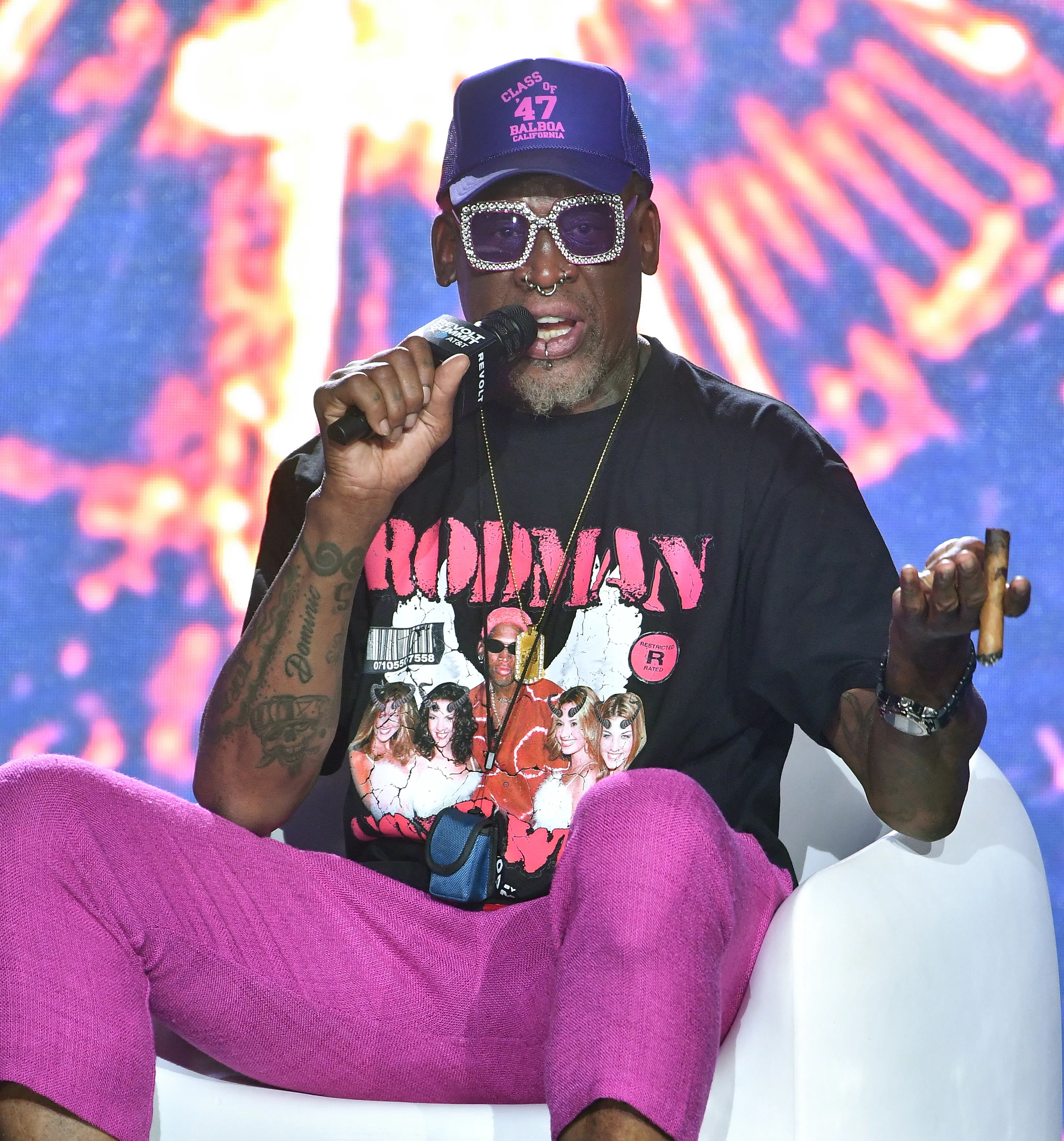 Dennis Rodman speaking on stage, wearing a purple hat and pink pants, holding a microphone in one hand and a cigar in the other.