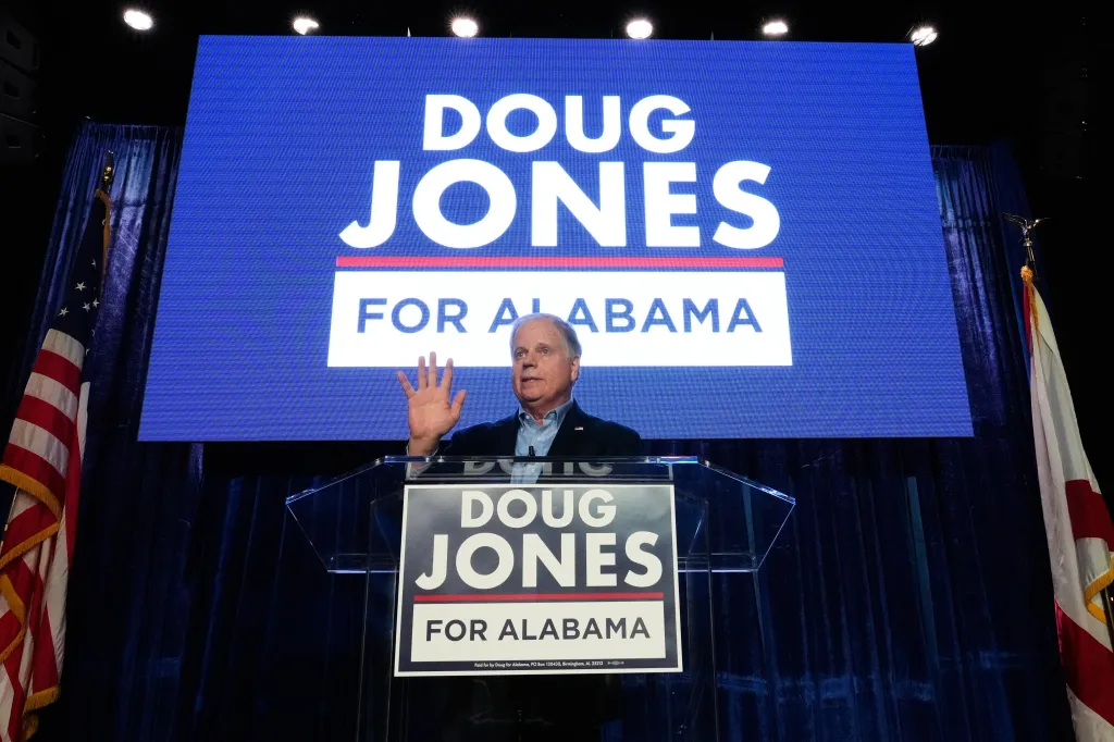Doug Jones speaking at an event, with "Doug Jones For Alabama" visible behind him on a screen and on the podium.