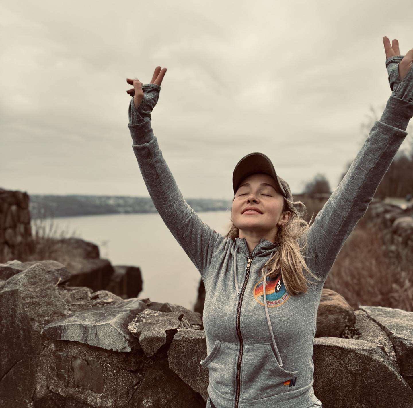 Hayden Panettiere with closed eyes and arms raised in front of a body of water.