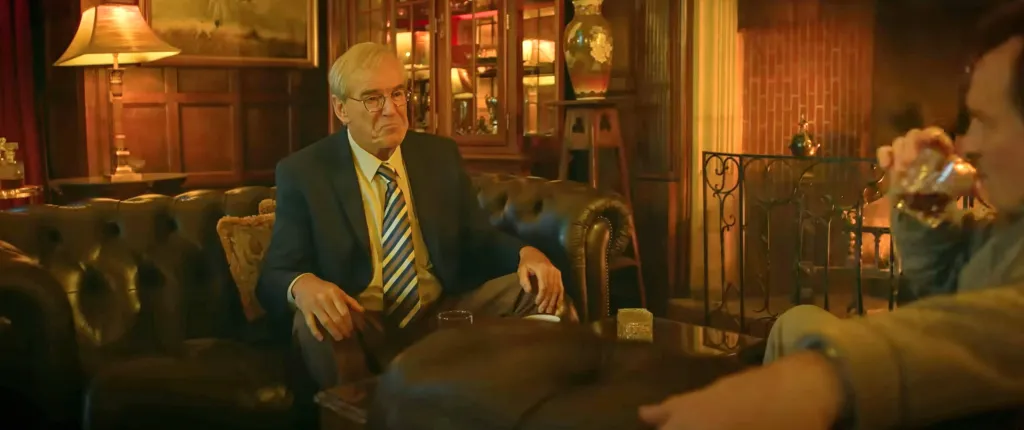 Pierce Brosnan in character as a man in a suit and tie sitting on a leather sofa.