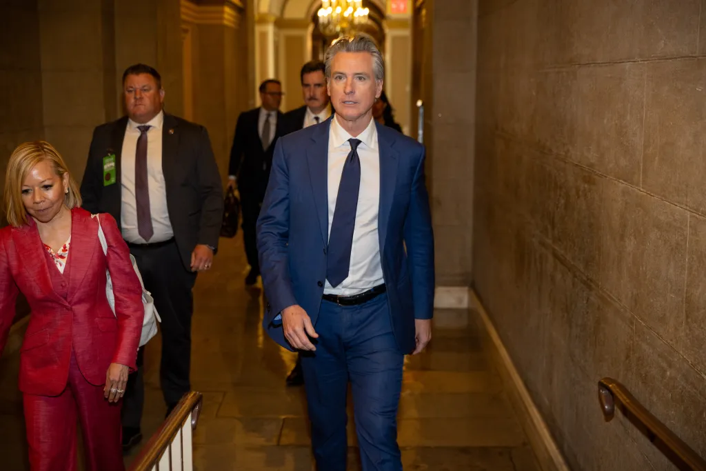 California Governor Gavin Newsom in a blue suit and tie walks through the halls of the U.S. Capitol, with a woman in a red pantsuit and security personnel.