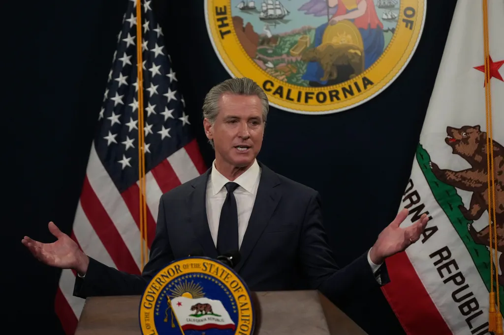 Governor Gavin Newsom speaking at a podium with flags behind him.