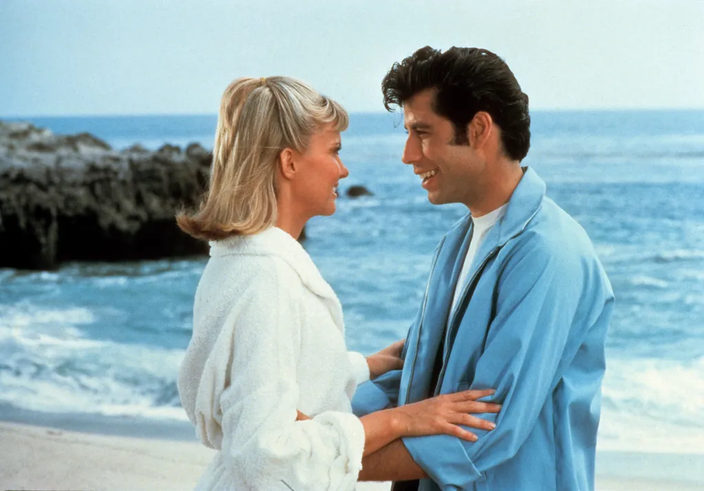 Olivia Newton-John and John Travolta smiling at each other on a beach.