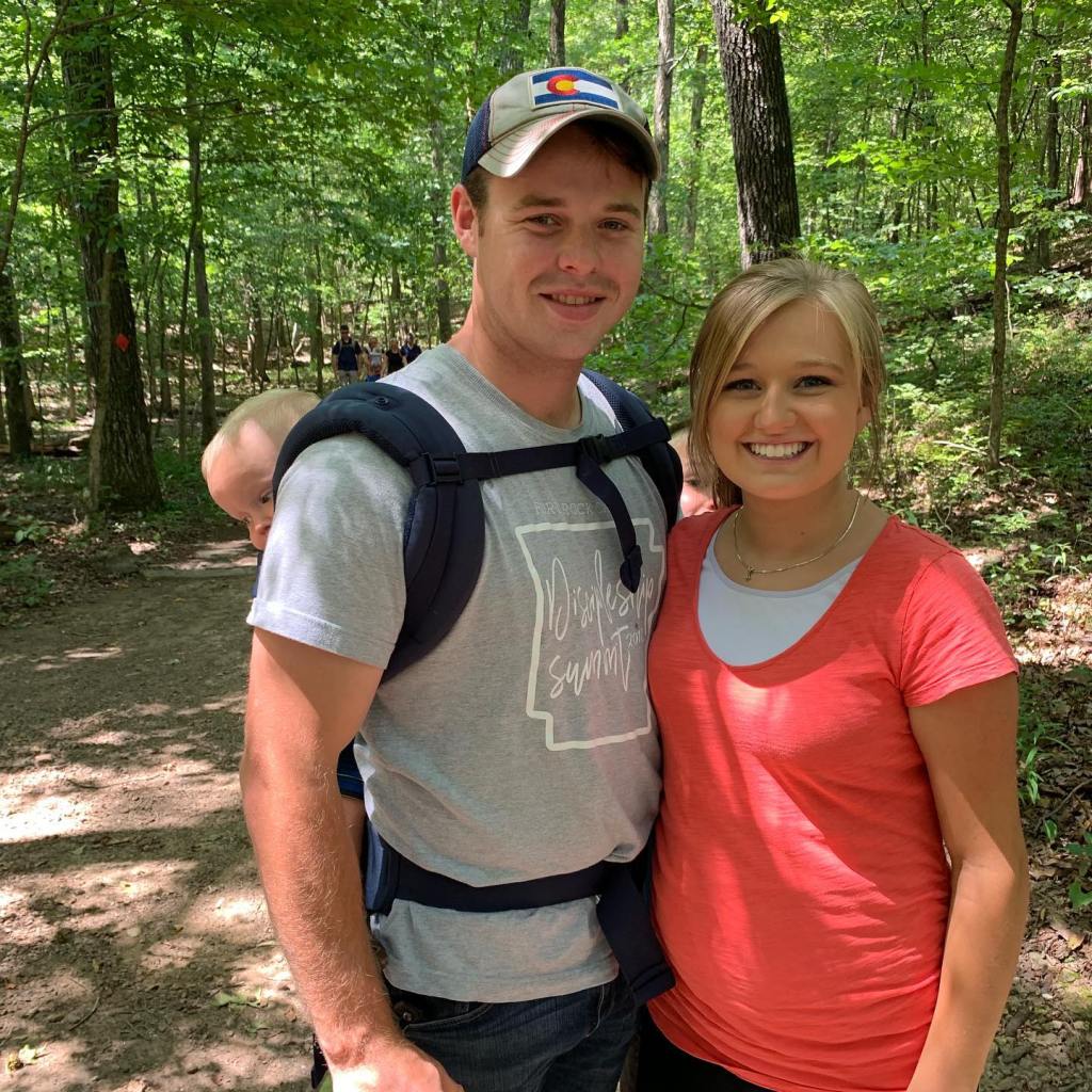 A man, woman, and baby in a carrier pose for a photo in a forest.