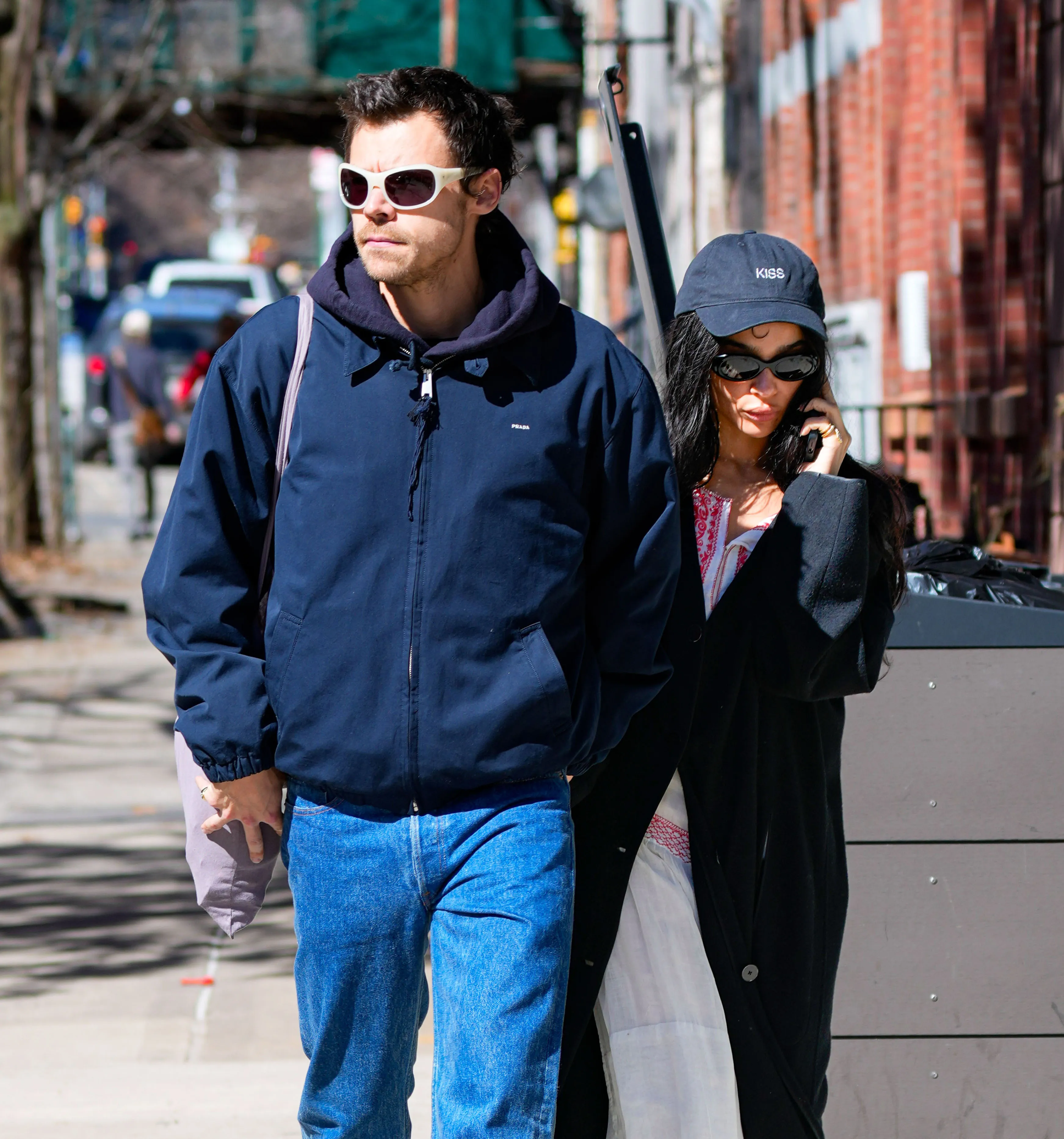 Harry Styles wearing a navy blue jacket and white sunglasses, walking next to Zoe Kravitz, who wears a black cap, black sunglasses, and a black coat.