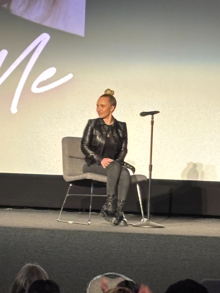 Hayden Panettiere sitting on a chair on a stage, facing right, with a microphone stand to her right.