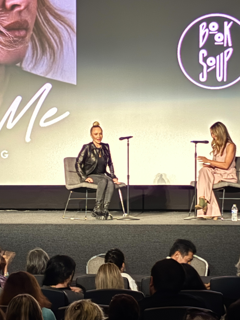 Hayden Panettiere speaking at an event, dressed in a black leather jacket and jeans, sitting next to another woman on a stage.