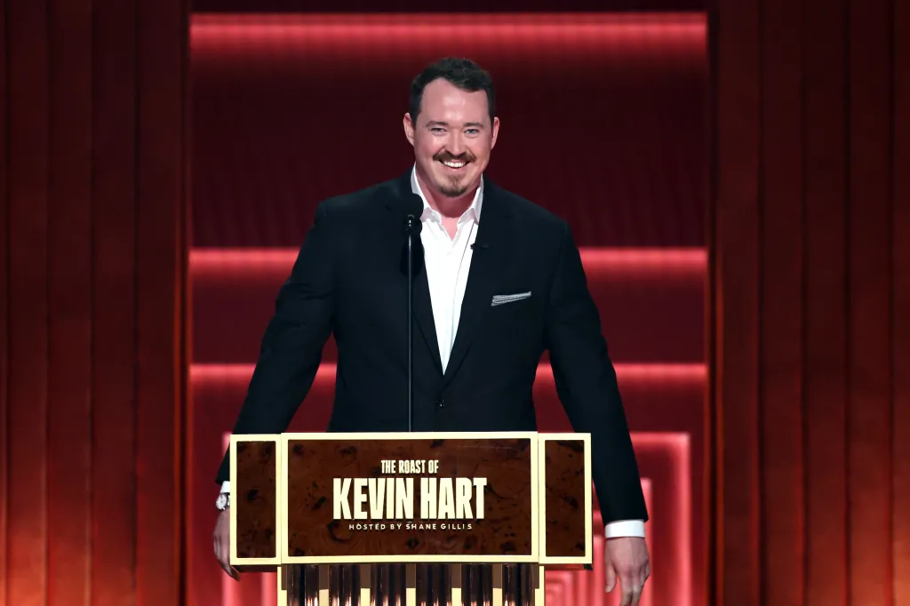 Shane Gillis at a podium labeled "The Roast of Kevin Hart" with a red lighted background.
