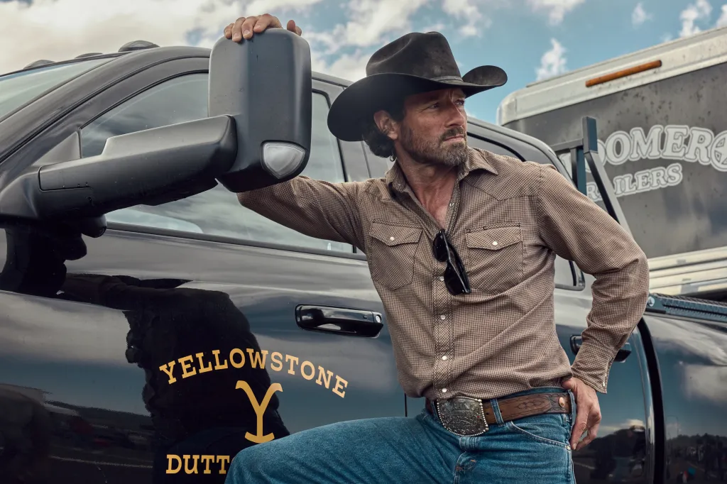 Ian Bohen as Ryan on Paramount Network's Yellowstone, wearing a cowboy hat, plaid shirt, jeans, and a belt with a large buckle, leaning on a black truck with the Yellowstone logo.