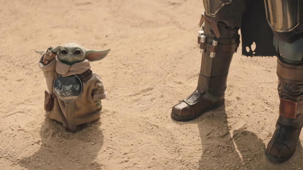 Grogu standing next to The Mandalorian's legs in a desert landscape.