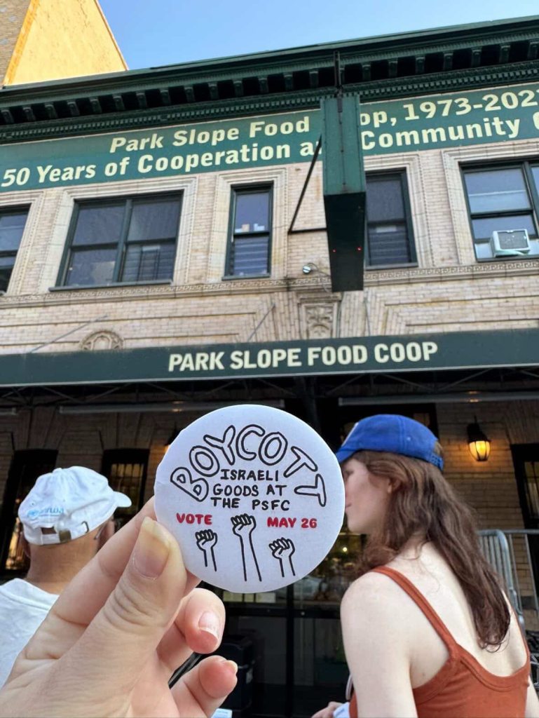 A hand holding a "Boycott Israeli Goods" button in front of the Park Slope Food Coop.