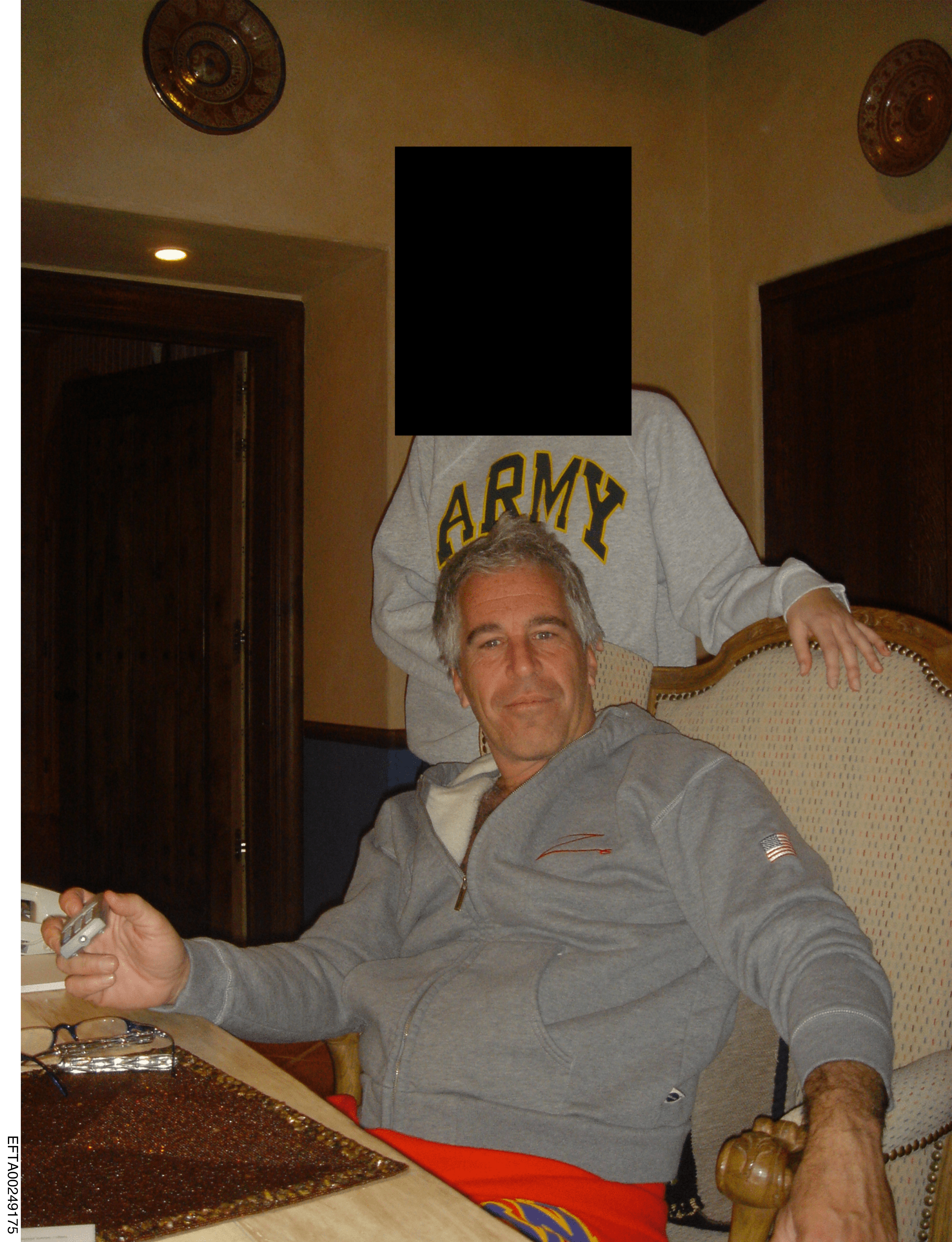 Jeffrey Epstein seated at a table, with another individual (face blacked out) standing behind him wearing an "ARMY" sweatshirt.