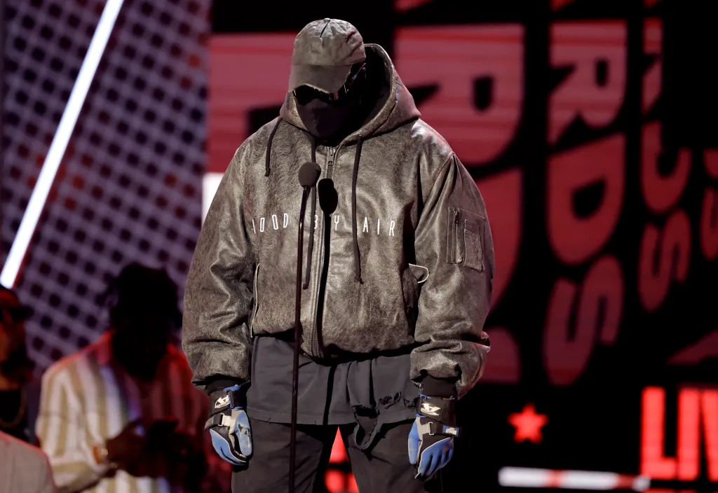 Kanye West on stage at the 2022 BET Awards wearing a black hooded outfit with a mask and sunglasses.
