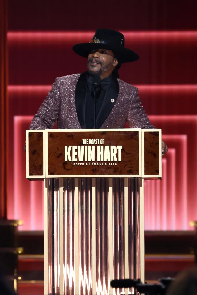 Katt Williams speaking at a podium with "The Roast of Kevin Hart" displayed, wearing a black hat and a shimmering suit.