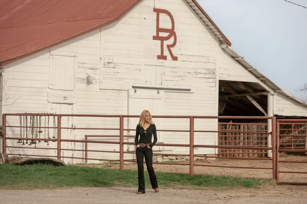 Kelly Reilly as Beth Dutton in black attire with hands in pockets standing in front of a white barn with a red "DR" logo.