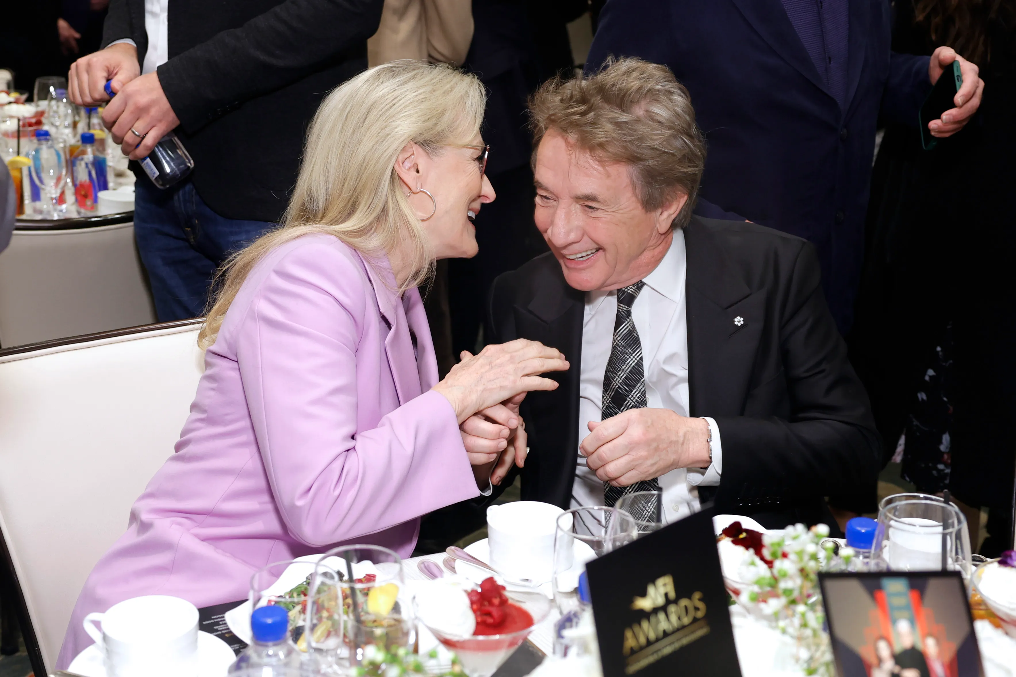 Meryl Streep and Martin Short laughing and holding hands at a table.