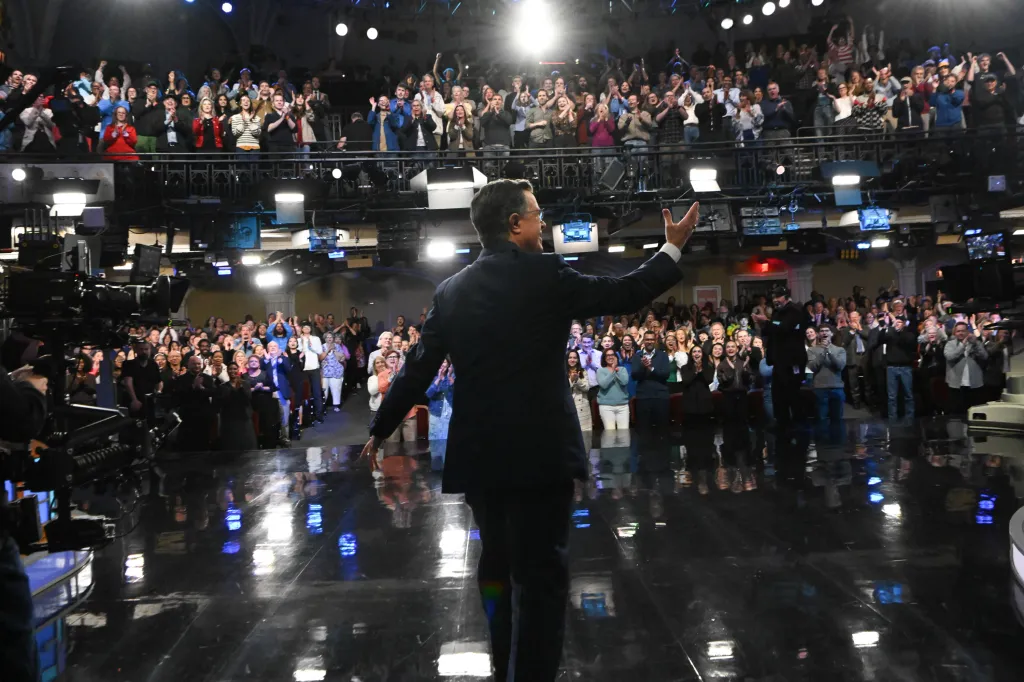 Stephen Colbert on stage, facing away from the camera, gesturing to an applauding audience.
