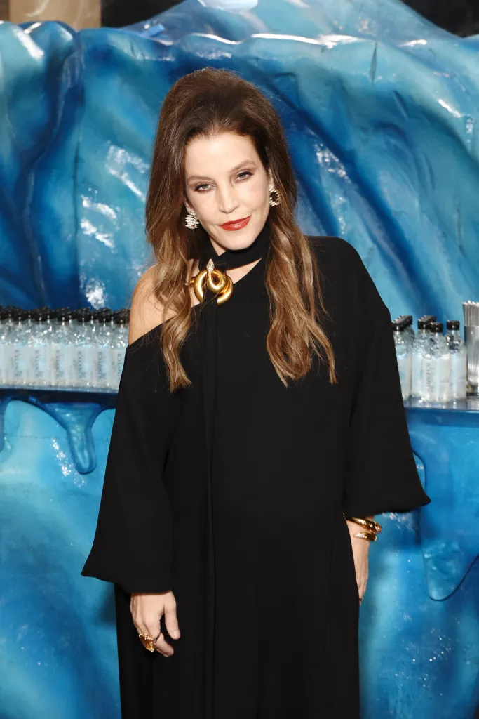 Lisa Marie Presley in a black dress with a gold necklace and earrings, standing in front of a blue ice-themed backdrop with Icelandic Glacial water bottles.