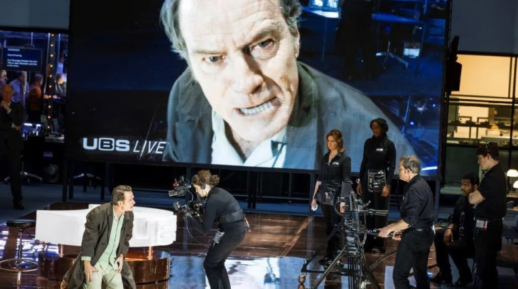 Bryan Cranston in "Network" on stage, with a large screen behind him showing his face, and camera operators in front.
