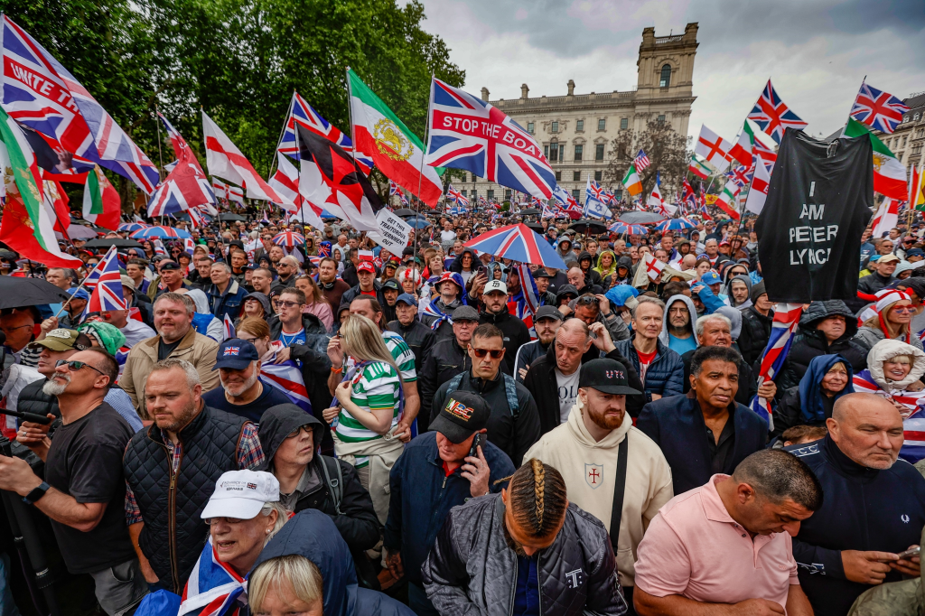 Crowds gathered at the Unite the Kingdom Rally in London, with many holding Union Jack flags, including one that reads "STOP THE BOAT" and another sign stating "GET THIS TRAITOROUS GOVERNMENT OUT."