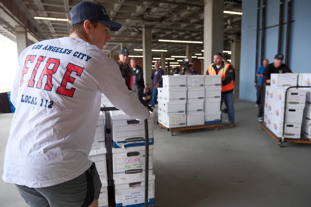 A Los Angeles firefighter wearing a "Los Angeles City Fire Local 112" shirt pushes a hand truck stacked with white Bankers Box storage boxes.