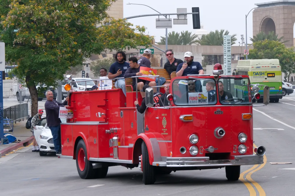 Firefighters on a vintage fire truck hold signs reading "Fund the Los Angeles Fire Department Now!" in a parade for a proposed ballot measure.