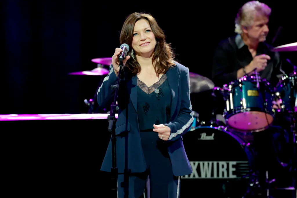 Martina McBride performing on stage, singing into a microphone with a drummer in the background.