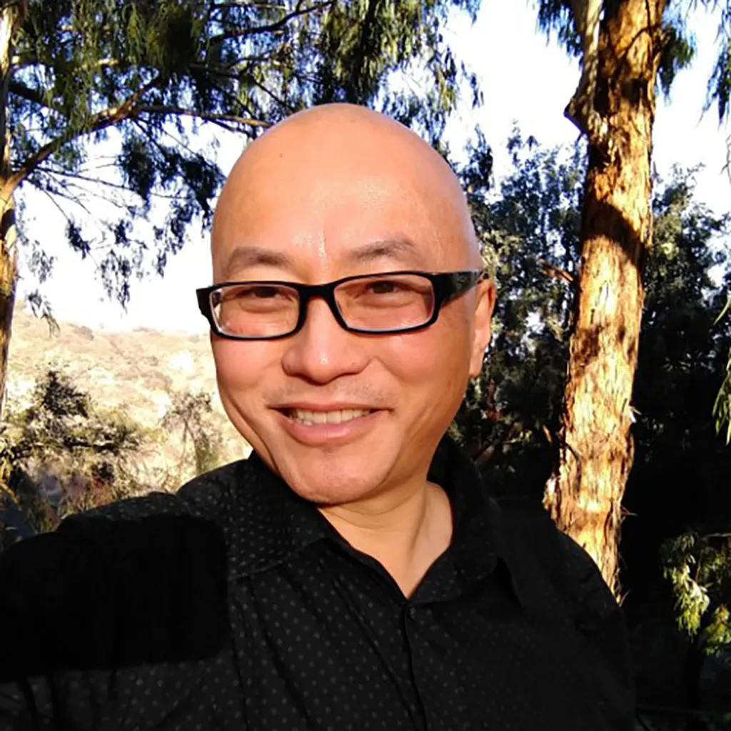 A man with glasses and a shaved head smiles at the camera, with trees and hills in the background.
