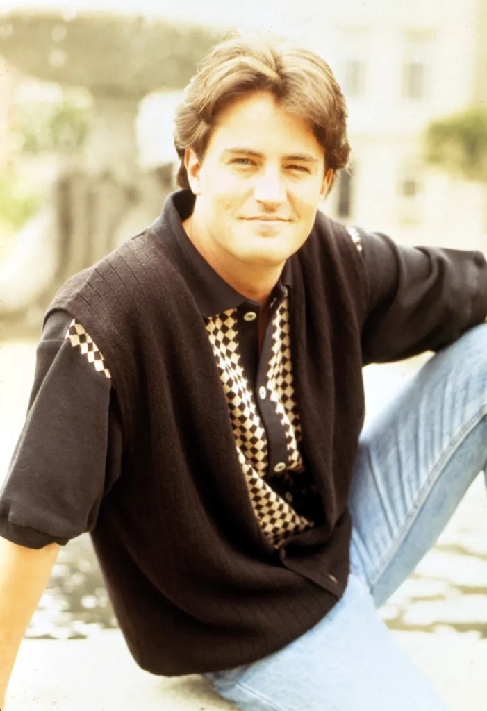 Matthew Perry in a black knitted polo shirt and light blue jeans, looking at the camera.