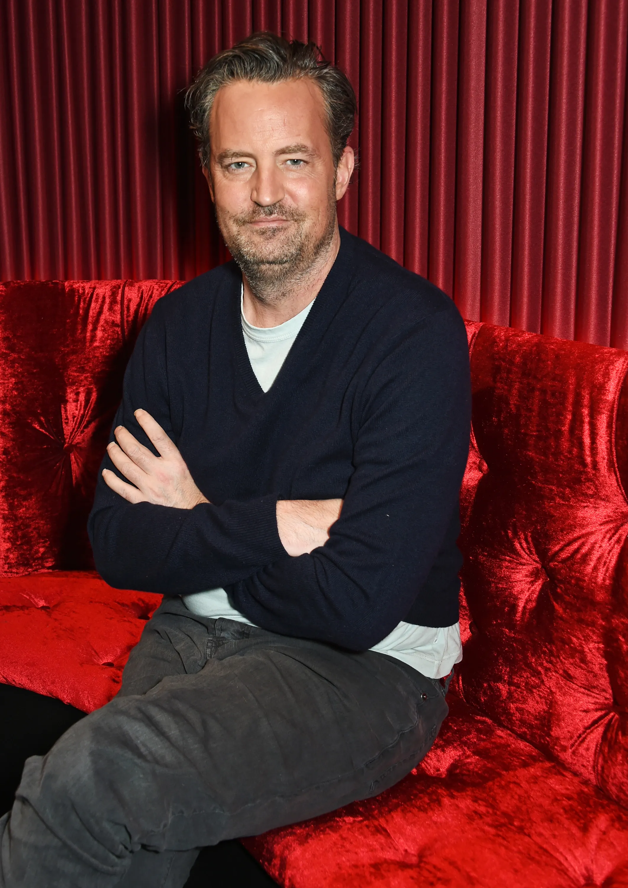 Matthew Perry posing for a photo on a red velvet couch.