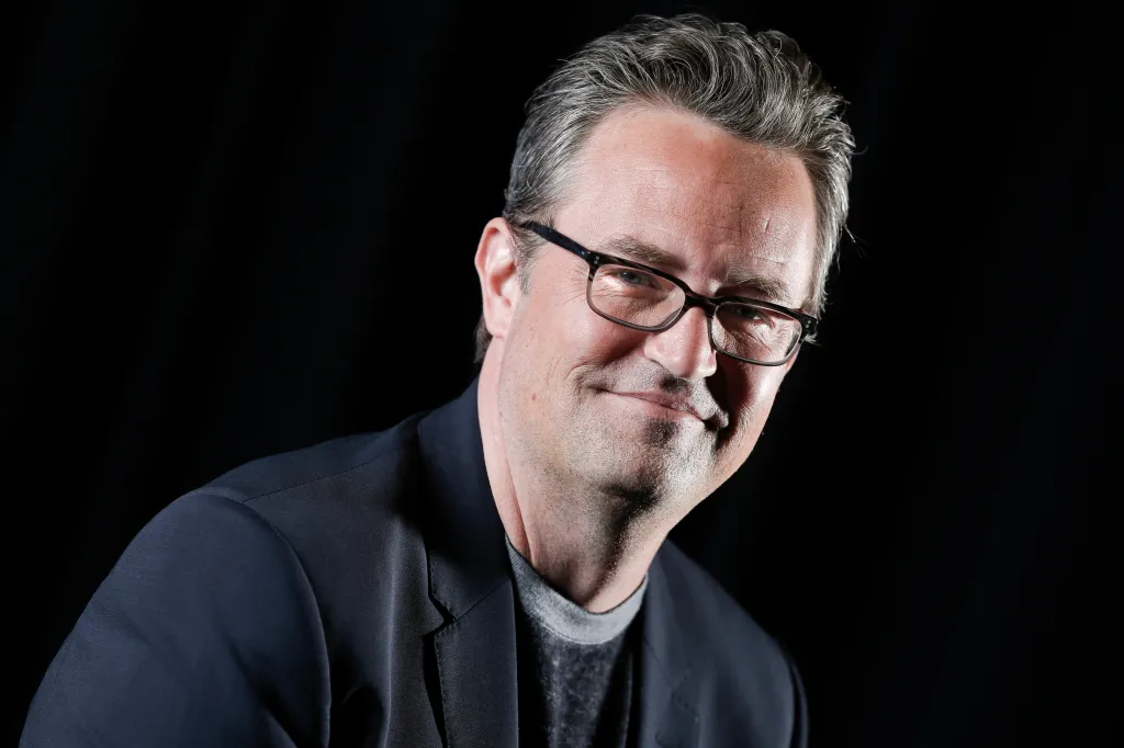 Matthew Perry, an actor, smiles wearing glasses and a dark blazer over a gray t-shirt.