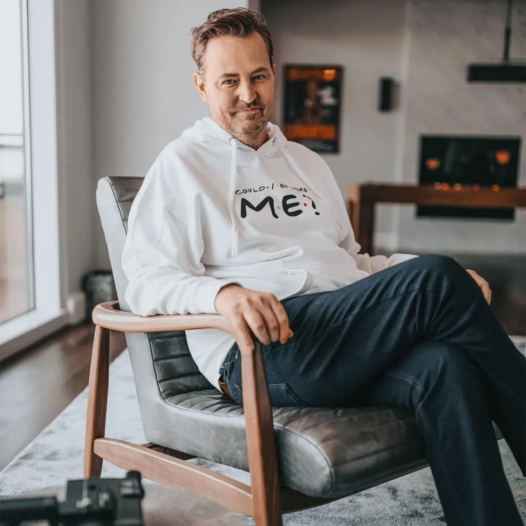 Matthew Perry sits smiling in a white hoodie that reads, "Could I be more ME?"
