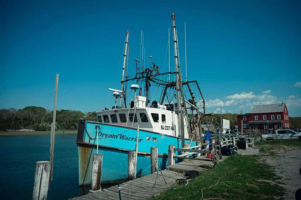 The boat "Dream Warrior" docked at Mattituck Inlet with a red building on the shore.